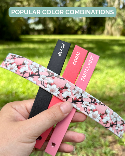 Hand holding color swatches with cherry blossom pattern, black, coral, and pastel pink labels against a blurred green outdoor background.