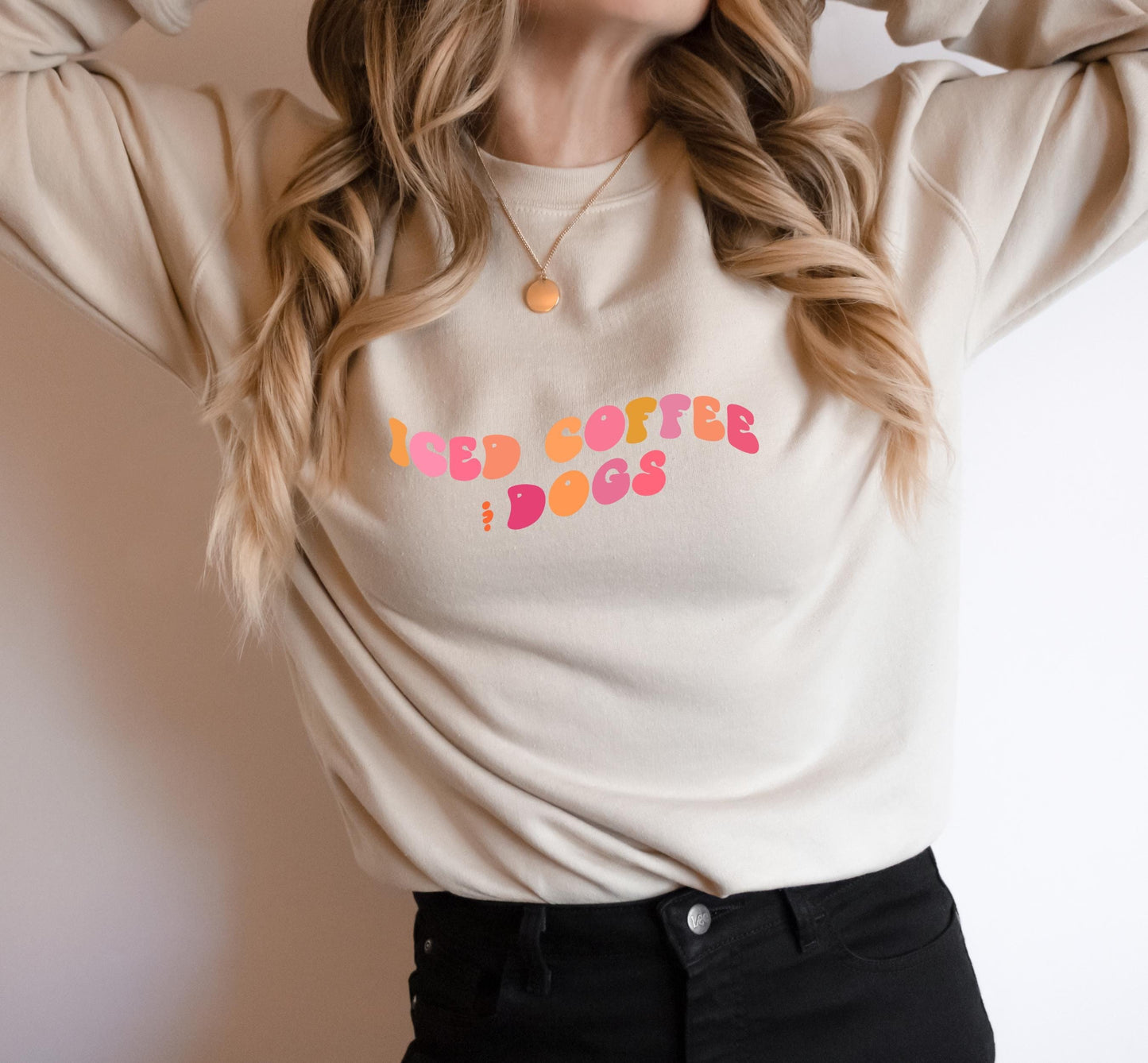 A person wearing a beige crewneck sweatshirt with the text 'iced coffee and dogs' printed in pink on the front.