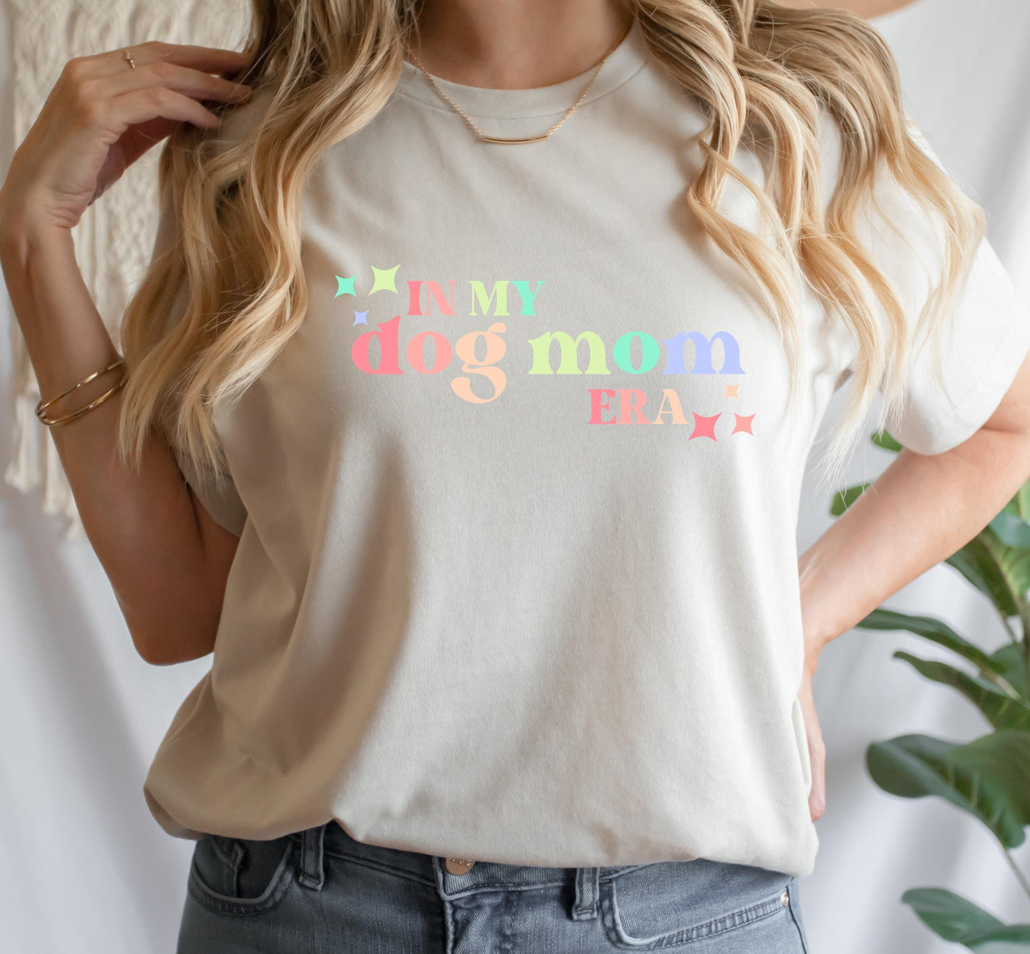 A person wearing a beige t-shirt with the text I'M MY DOG MOM ERA printed on the front, paired with blue jeans.