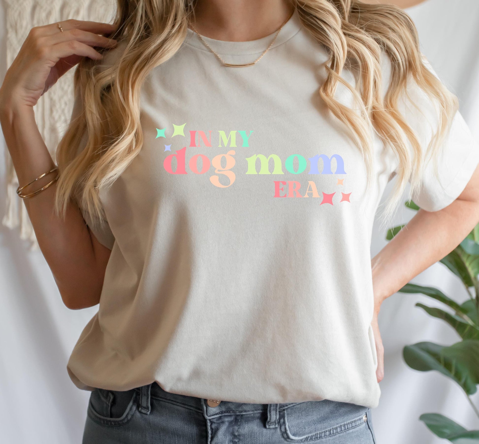 A person wearing a beige t-shirt with the text I'M MY DOG MOM ERA printed on the front, paired with blue jeans.