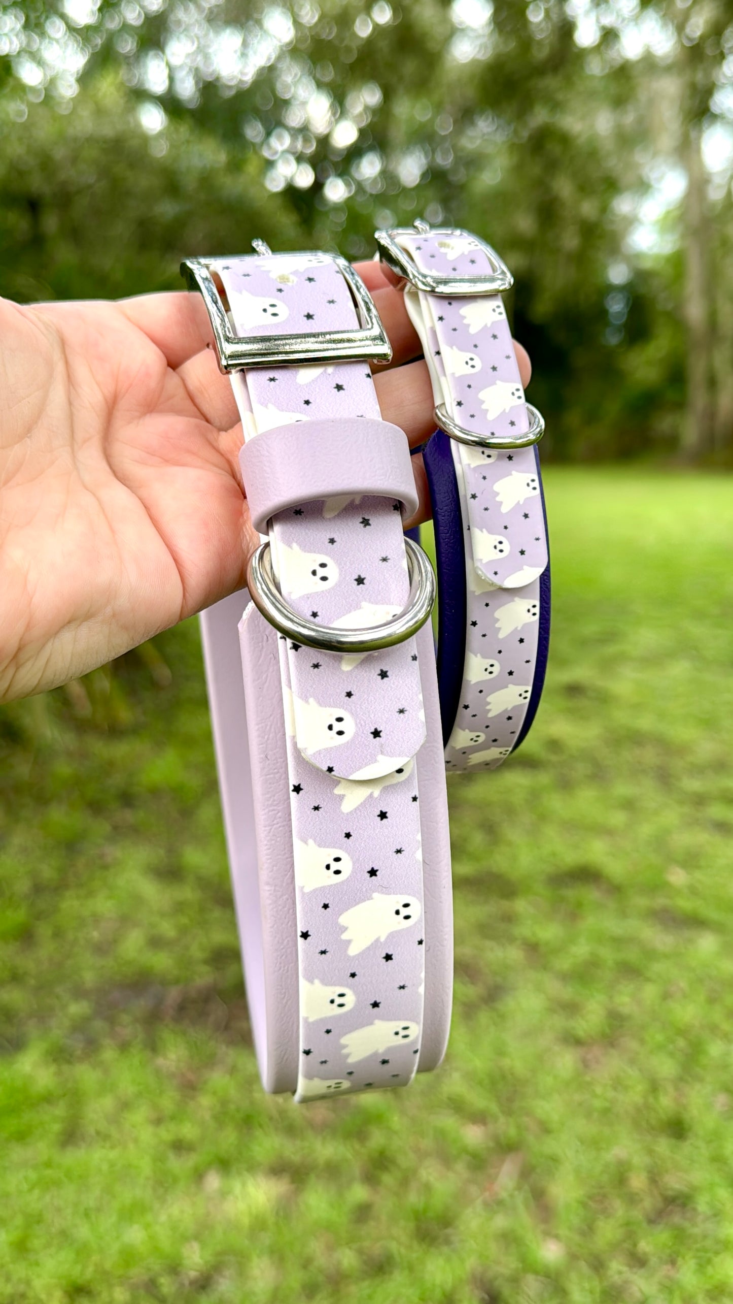 Two purple dog collars with ghosts and star patterns held by a hand against a green outdoor background.