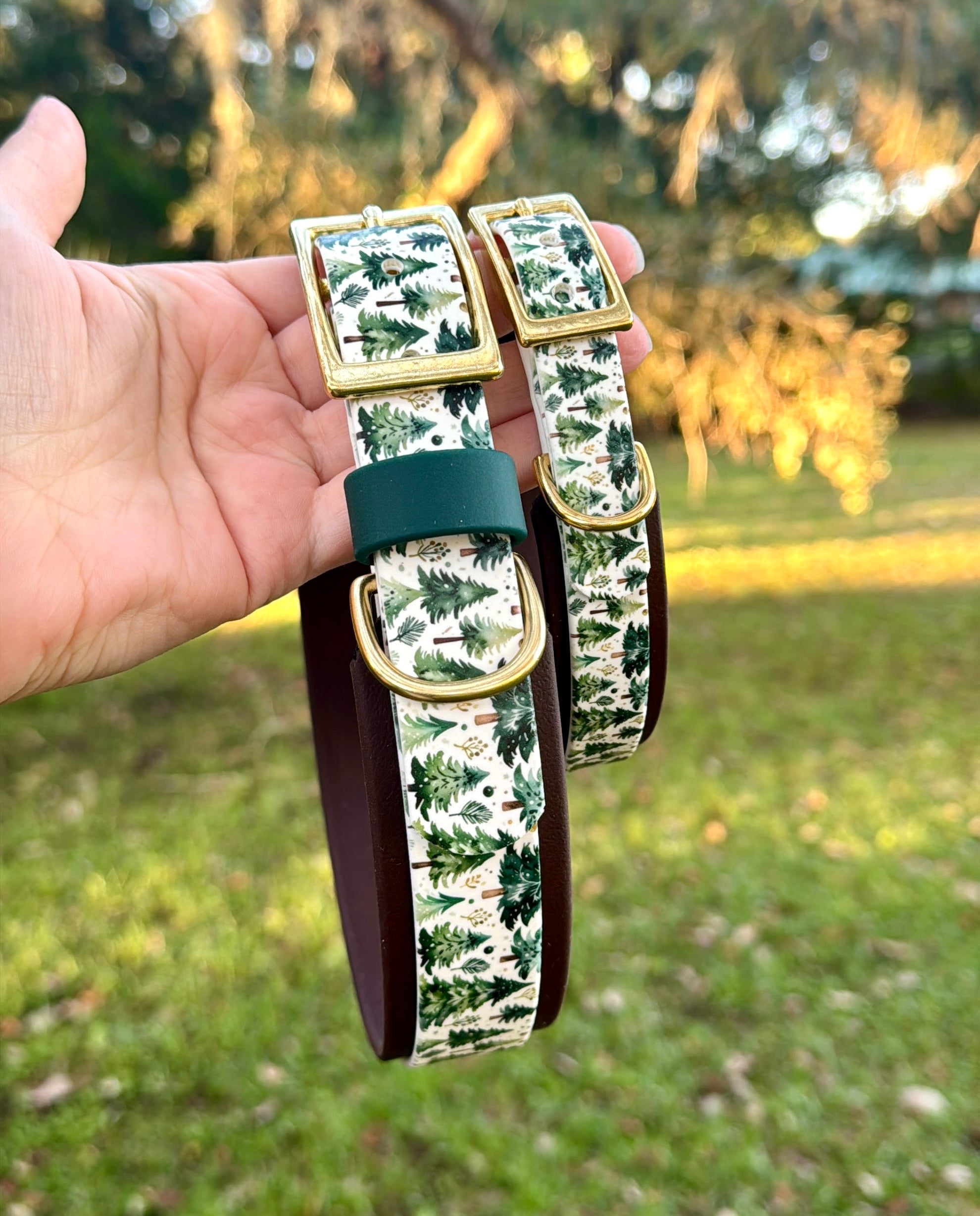 Two dog collars with winter trees that are green and gold held by a hand against a blurred natural background.